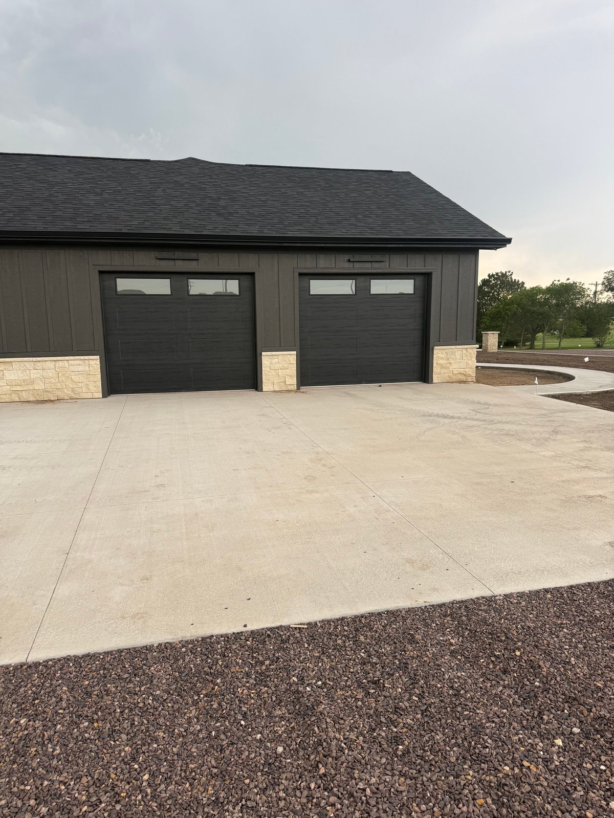 Modern dark gray garage with two black sectional doors, attached to a tan metal building with concrete driveway and gravel border