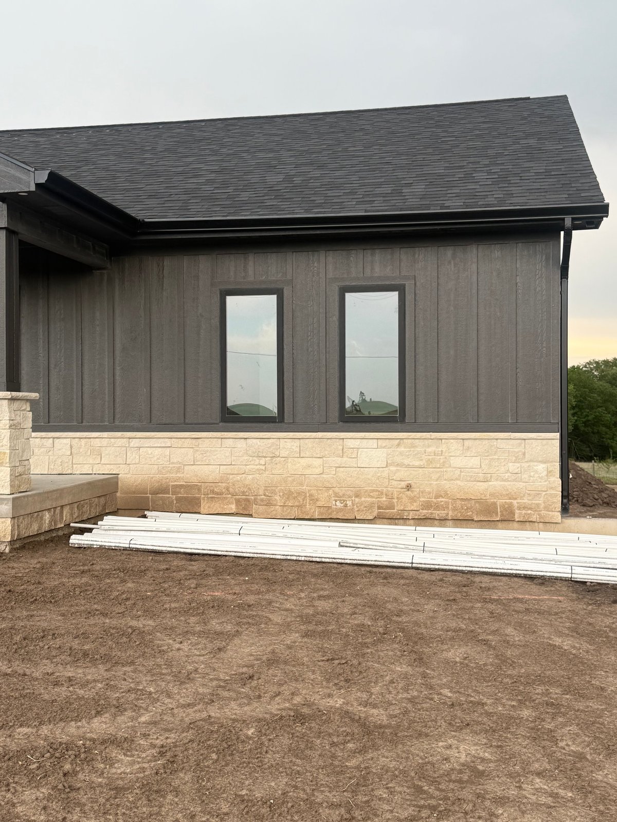 Modern residential home with dark gray siding, stone foundation, two rectangular windows, and pitched roof under construction on bare ground