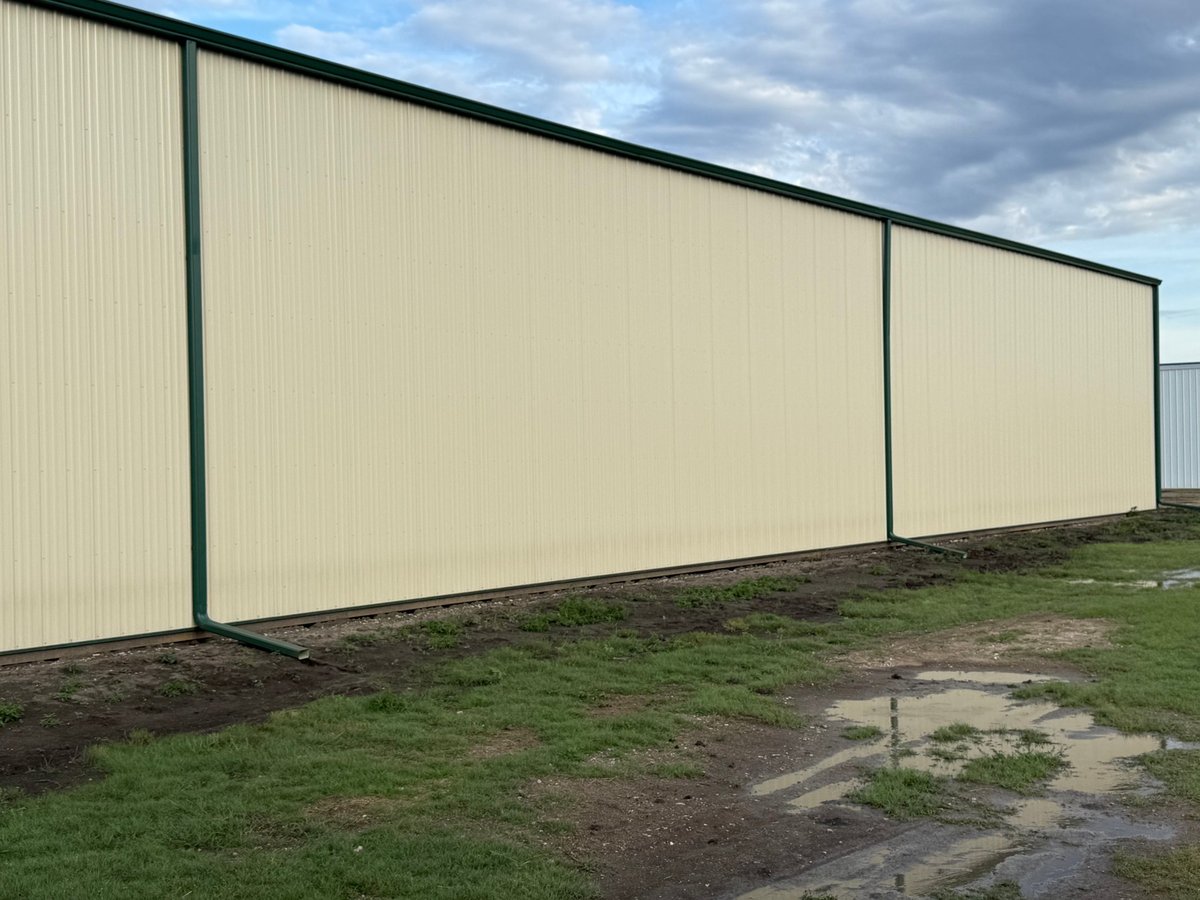 Large beige corrugated metal agricultural building with black frame and doors alongside grassy field with dirt path