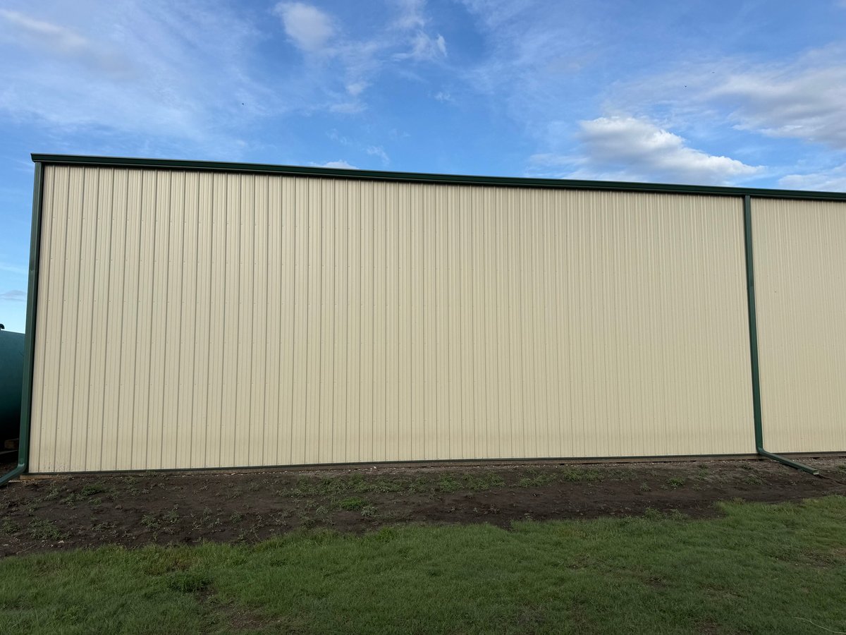Large corrugated metal industrial building with cream-colored siding and dark roof against blue sky with green grass in foreground