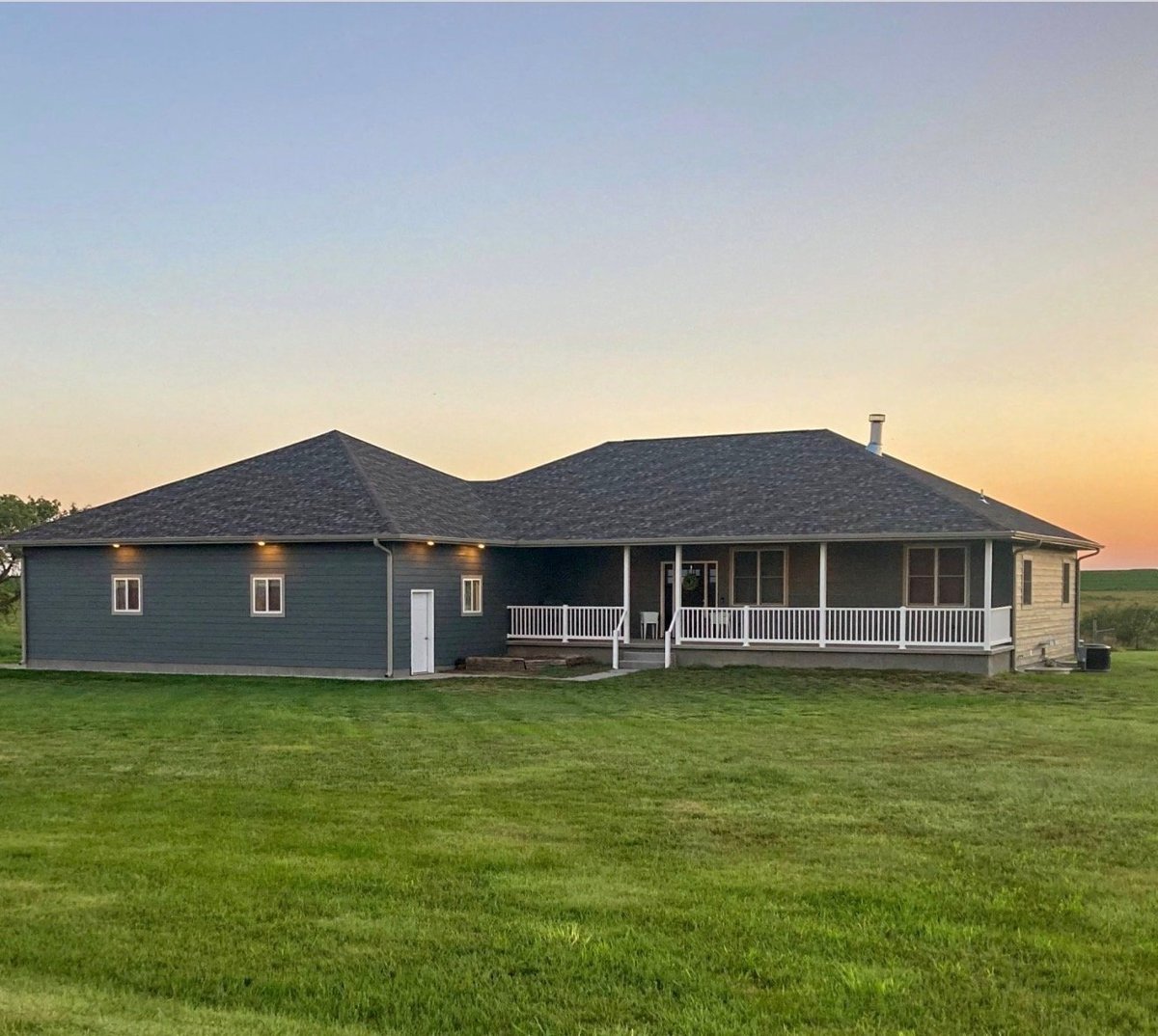 Modern gray residential house with wrap-around porch at sunset, set on expansive green lawn