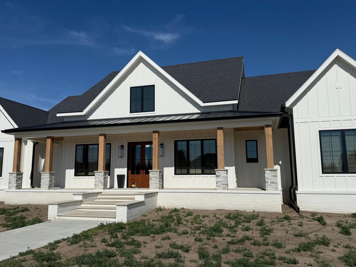 Modern white farmhouse with black roof, wooden columns, front porch, and landscaped yard under clear blue sky